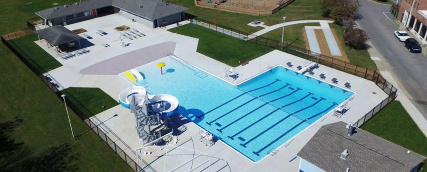 Aerial view of an outdoor swimming pool complex with a lap pool, water slides, splash features, chairs, and surrounding buildings, all enclosed by a fence with pathways and green lawn areas nearby.