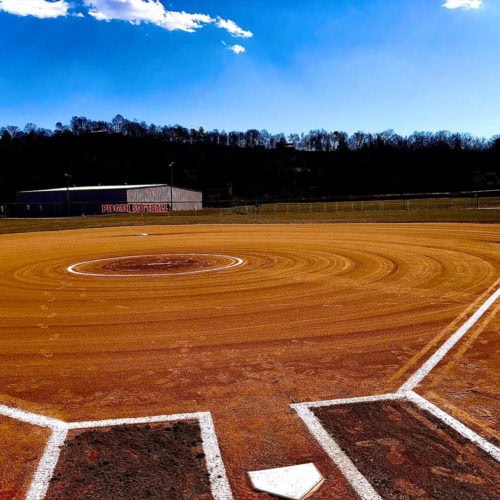 A well-maintained softball or baseball field is seen from behind home plate on a sunny day, with clear sky and trees in the background.