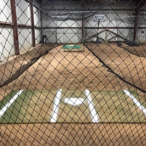 Indoor baseball or softball batting cage with turf mats, white batters box lines, dirt floor, and netting enclosing the area. A pitching mound and a basketball hoop are visible in the background.