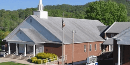 A brick church with white trim, a steeple, and steps leading to the entrance. An American flag is on a flagpole in front, with trees and green hills in the background.