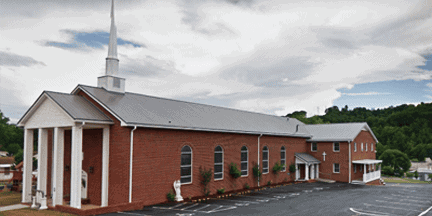 A large red-brick church with white columns, arched windows, and a tall white steeple, sits beside a parking lot under a cloudy sky.