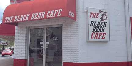The entrance of The Black Bear Cafe with a red awning and a sign featuring a bears face. The building is white with red trim. A car is visible through the glass door.