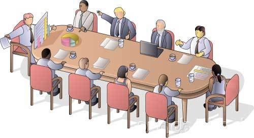 Nine people sit around a large oval conference table with documents, coffee cups, a laptop, and charts, engaged in a meeting or discussion.