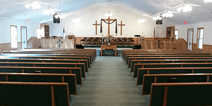 Interior of a church with empty green pews, a carpeted aisle, wooden podium, and three crosses at the front. Ceiling fans and lights hang above, and American flags stand near the stage.