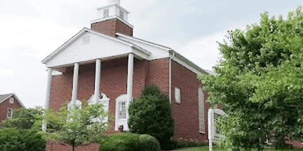 A red brick church with white columns at the entrance, a small steeple, and green trees around the building under a cloudy sky.