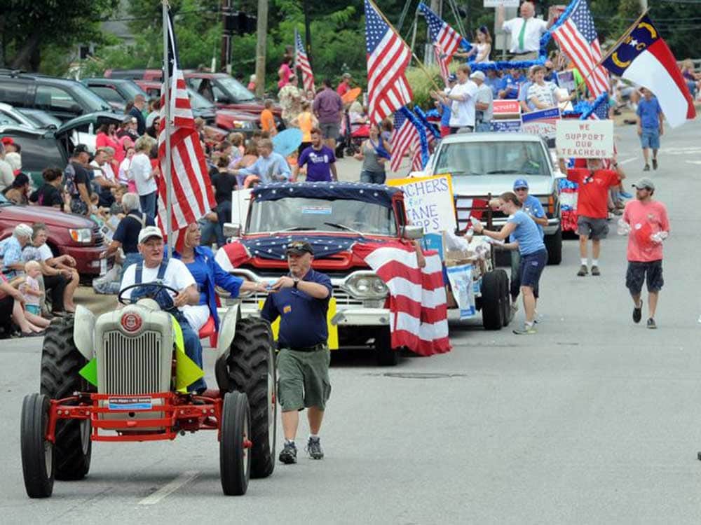 A parade with people waving American flags, walking and riding on decorated vehicles. Spectators sit along the street, and signs supporting teachers are visible.