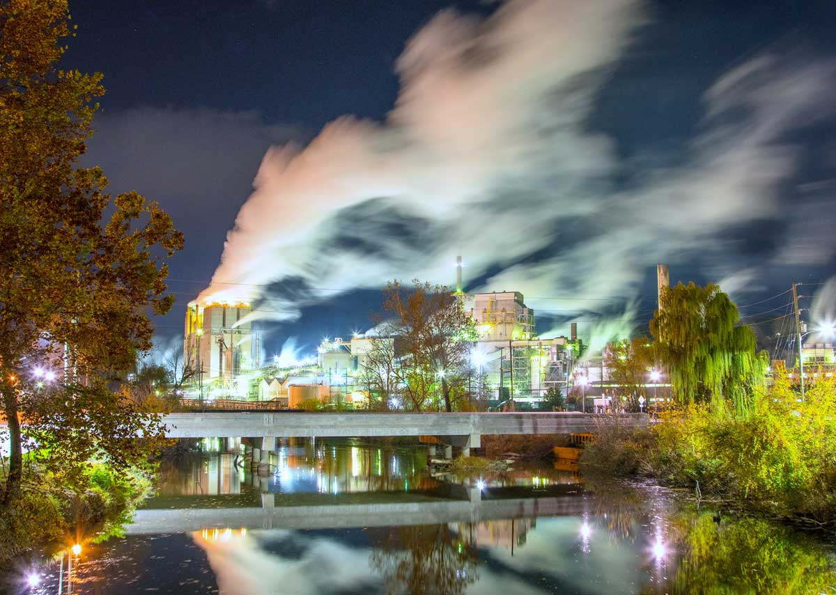 A brightly lit industrial factory emits large plumes of steam or smoke into the night sky, with trees and a river reflecting the scene in the foreground.