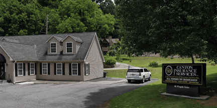 A beige building with dormer windows sits beside a driveway, with a white SUV parked outside. A black and gold sign reads Canton Insurance Services. Green trees and grass are in the background.