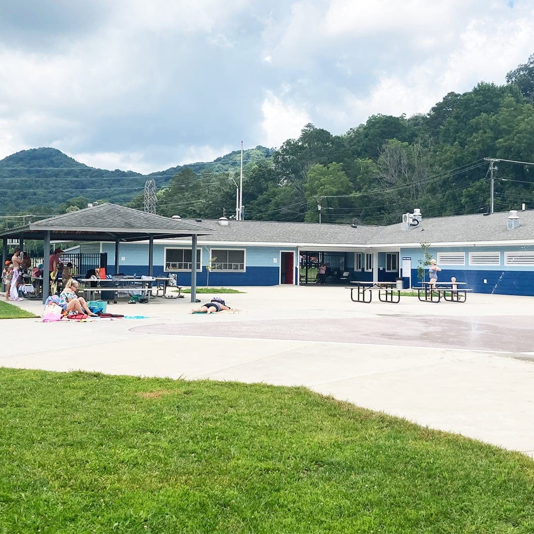 People relax on towels and benches near a blue and white community building, with mountains and trees in the background under a partly cloudy sky.