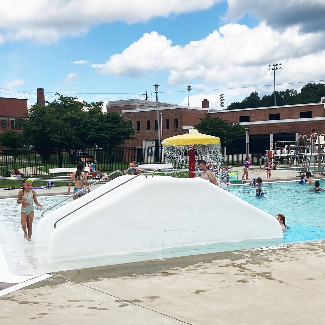 Children play in an outdoor public pool with a small white slide and a yellow mushroom-shaped water feature. Other people swim and relax nearby. The sky is partly cloudy, and buildings are visible in the background.