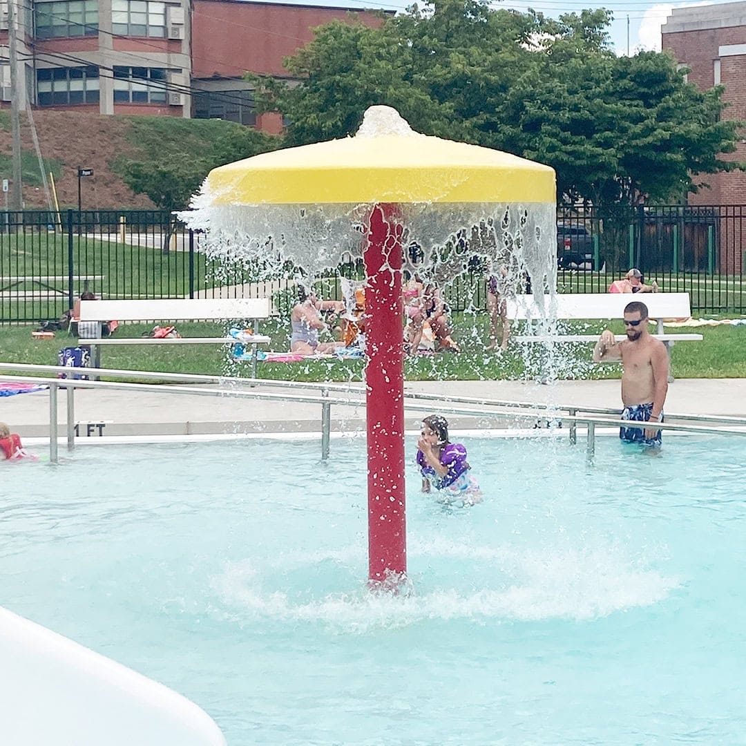 A splash pad features a yellow mushroom-shaped fountain with water cascading down. Children play nearby while adults watch from lounge chairs in the background. Outdoor pool area on a sunny day.