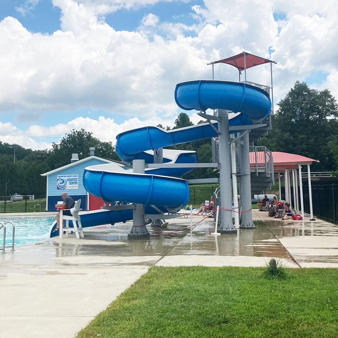 A large blue water slide curves into an outdoor swimming pool. Children wait in line while a lifeguard sits nearby. The sky is partly cloudy, and there are trees and buildings in the background.