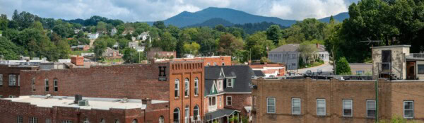 A small cityscape with brick buildings in the foreground, lush green trees, and rolling mountains under a partly cloudy sky in the background.