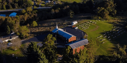 Aerial view of a church surrounded by trees, with a cemetery on one side and a few vehicles parked nearby under a clear sky.