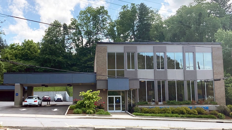 A modern two-story office building with large windows, surrounded by trees. Several parked cars are visible near the entrance, and the sky is partly cloudy.