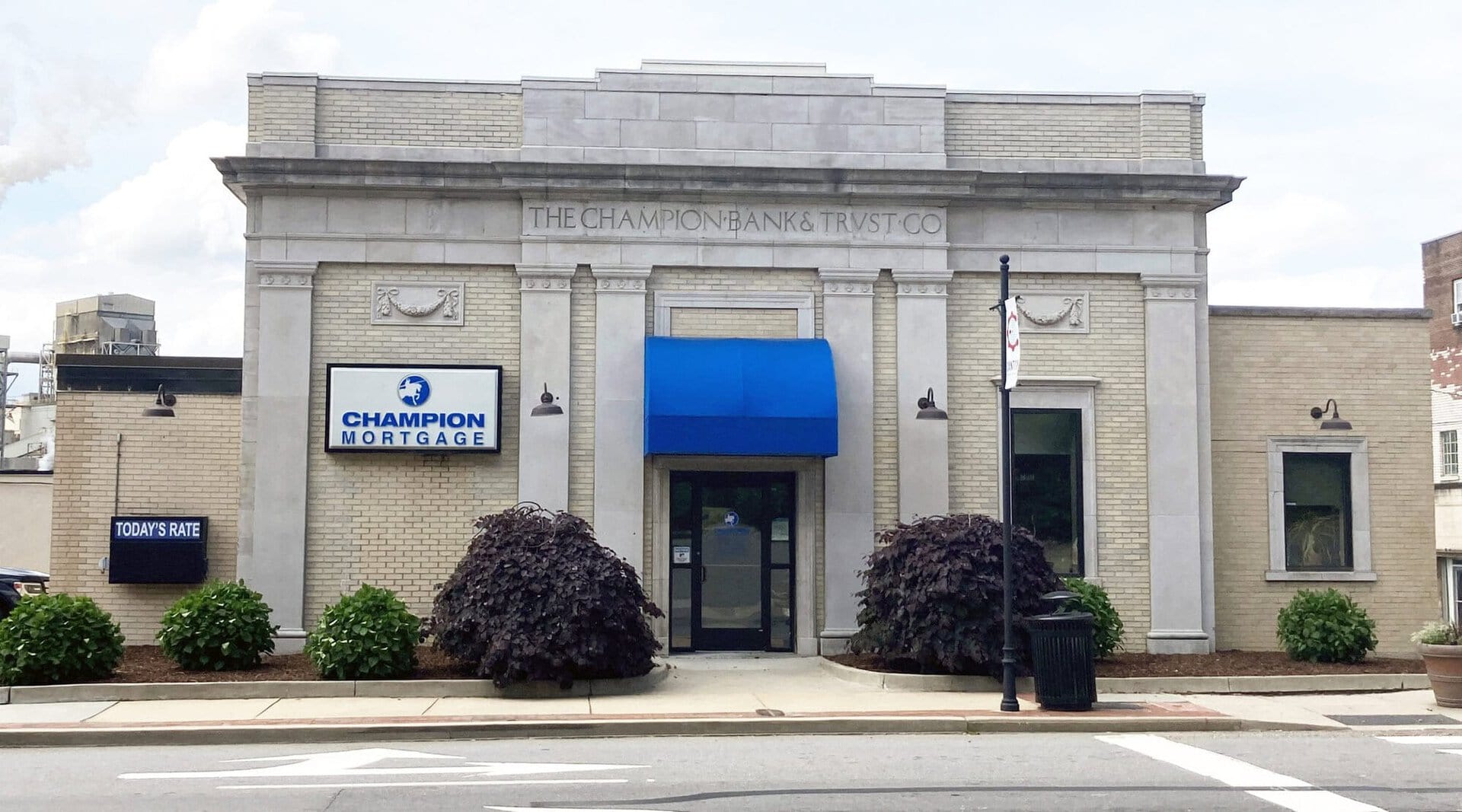 A tan brick bank building with columns, a blue awning, Champion Mortgage signs, and landscaped bushes in front. A small digital sign displays TODAYS RATE. The street is visible in the foreground.