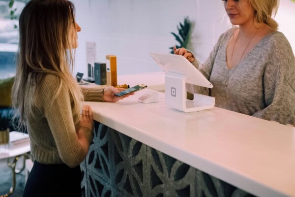 Two women stand at a counter; one is holding a phone, appearing to make a payment, while the other operates a tablet-style register. The scene is in a modern, well-lit retail or service space.
