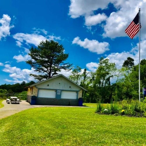 A small building with a closed garage sits near a paved path, surrounded by green grass and trees. An American flag flies on a pole under a bright blue sky with fluffy clouds.
