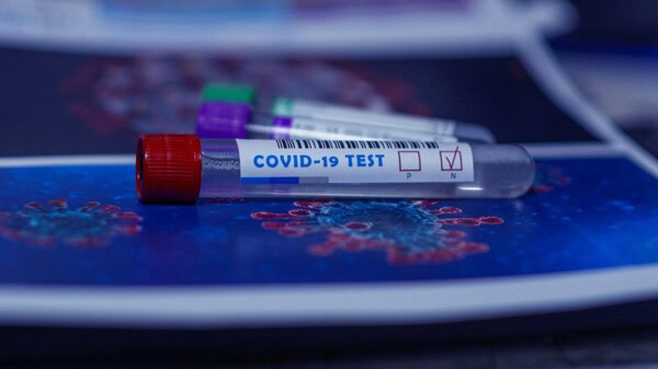 A COVID-19 test tube with a red cap lies on a surface, labeled with a positive test result. Blurred images of the coronavirus are visible in the background.