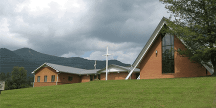 A brick church with a large window and white cross stands on a grassy hill, with mountains and cloudy skies in the background.