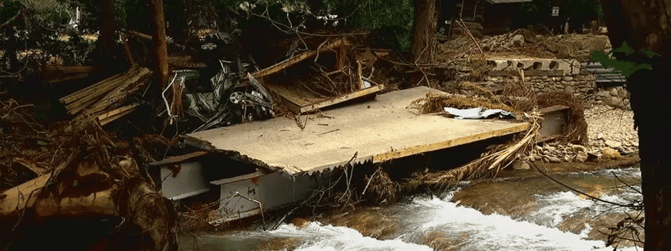 A collapsed concrete bridge lies in a creek, surrounded by debris and fallen trees after a flood, with muddy water flowing beneath the wreckage.
