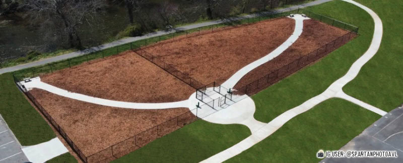 Aerial view of a fenced dog park with two large separate sections, mulched ground, benches, and curved concrete pathways, surrounded by green grass.