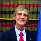 Smiling person with glasses and light brown hair wearing a suit, red tie, and pin, standing in front of a bookshelf filled with legal books and partially visible blue flag.
