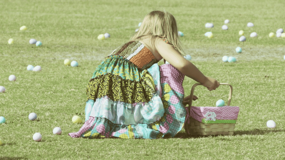 A young girl in a colorful dress gathers Easter eggs into a basket on a grassy field scattered with plastic eggs.