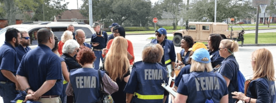 A group of FEMA workers, wearing blue vests with FEMA on the back, gather in a circle outdoors, listening to a person in red, with vehicles and trees in the background.