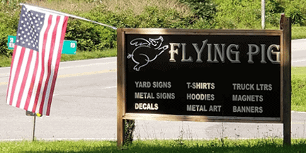 Sign for FLYING PIG with a flying pig illustration, listing items like yard signs, decals, T-shirts, hoodies, and more. An American flag is displayed beside the sign near a roadside.