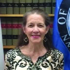 A woman with long brown hair wearing a patterned top and earrings stands in front of bookshelves and a blue flag, smiling at the camera.
