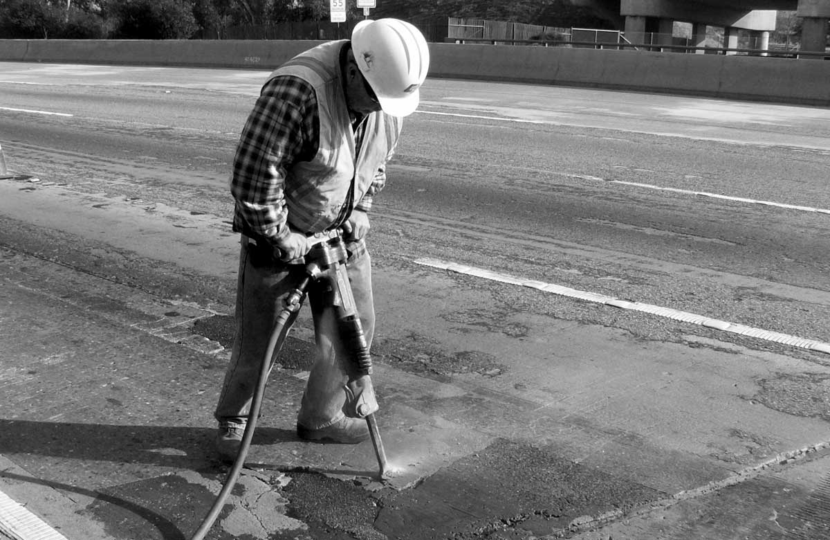 A construction worker in a hard hat and vest uses a jackhammer to break up pavement on a road. The scene is in black and white.