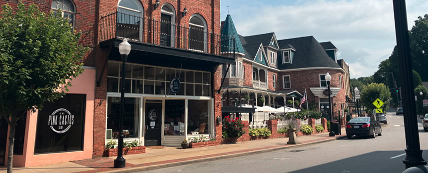 A quiet street scene in a small town with brick buildings, storefronts, outdoor seating, a tree, parked cars, and a streetlamp under a partly cloudy sky.