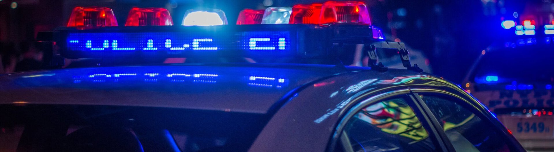 Close-up of a police cars flashing lights at night, with the word POLICE illuminated in blue. Another police vehicle with lights is visible in the blurred background.