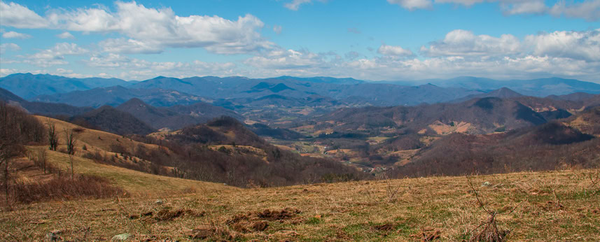 Rolling hills and mountains under a partly cloudy sky, with patches of green and brown vegetation, stretch into the distance in this scenic landscape.