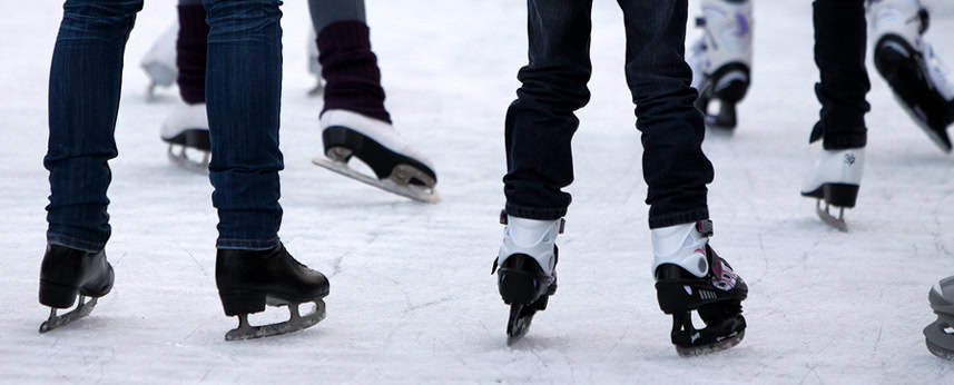 Several people wearing ice skates glide on an ice rink. Only their legs and skates are visible, with some moving forward and others standing still.