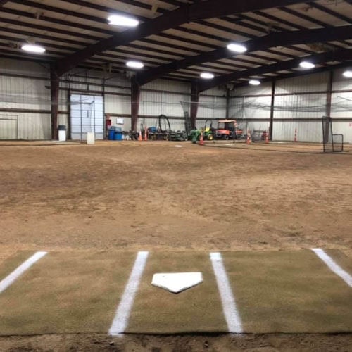 Indoor baseball facility with a home plate and batters box marked on turf. The floor is mostly dirt, and equipment is visible in the background under a metal roof. Bright lights hang from the ceiling.