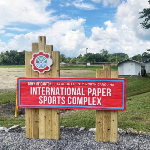 A red sign reads International Paper Sports Complex, Town of Canton, Haywood County, North Carolina, mounted on wooden posts outdoors with grass and a small building in the background.