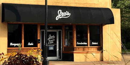 A restaurant with black awning displaying Jros. Windows have white text listing menu items: burgers, subs, sandwiches, and salads. The building is tan with large windows and a glass door. Plants are visible in front.