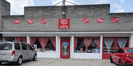 Storefront with red and white decor, red curtains in large windows, a red door, and a red sign above the entrance. Two vehicles are parked in front of the building on a concrete lot under a partly cloudy sky.