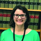 A woman with short brown hair and glasses, wearing a bright green top and a black necklace, smiles in front of bookshelves filled with law books.