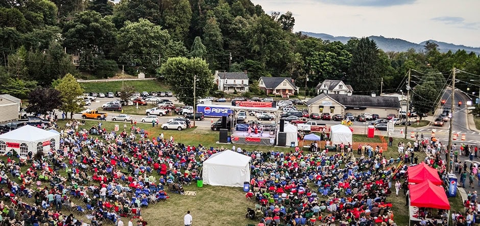 A large crowd sits on a grassy field at an outdoor event with tents, a stage, and parked cars nearby. Trees and houses border the area, with hills visible in the background under a cloudy sky.