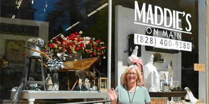 A woman smiles and waves in front of a store called Maddies on Main, which has a window display featuring flowers, decor, and vintage items.