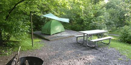 A green tent sits on a wooden platform with a tarp overhead. Nearby, theres a picnic table and benches on a gravel area, surrounded by lush green trees. A fire pit is visible in the foreground.