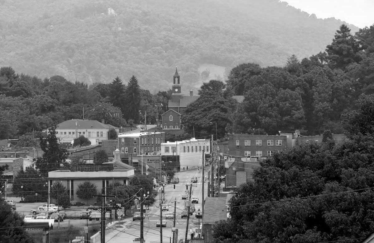 A black and white photo of a small town with buildings, cars on the street, trees, and a church with a steeple. Hills covered in trees rise in the background.