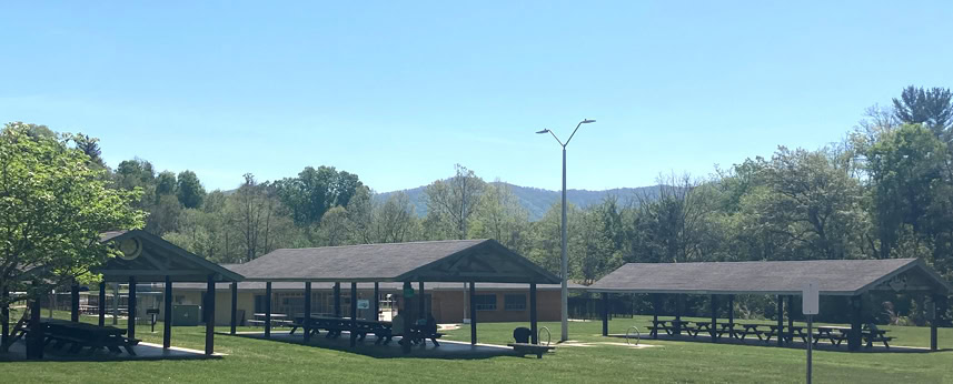 Three picnic shelters with tables stand on a grassy field surrounded by trees and distant mountains under a clear blue sky.