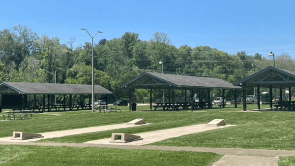 Open grassy park area with three covered picnic pavilions, several picnic tables, and a few cornhole boards on concrete pads. Trees and parked cars are visible in the background under a clear blue sky.