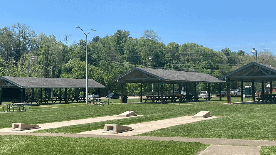 Open grassy park area with three covered picnic pavilions, several picnic tables, and a few cornhole boards on concrete pads. Trees and parked cars are visible in the background under a clear blue sky.