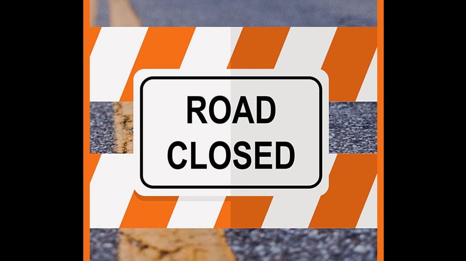A Road Closed sign with orange and white stripes blocks a road, indicating the street is not accessible to vehicles.