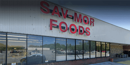 Exterior of a SAV-MOR FOODS grocery store with large red letters on the building and glass windows reflecting parked cars and trees.